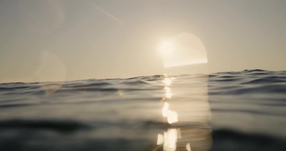 From a water level perspective silhouetted surfers ride waves towards the bright rising sun