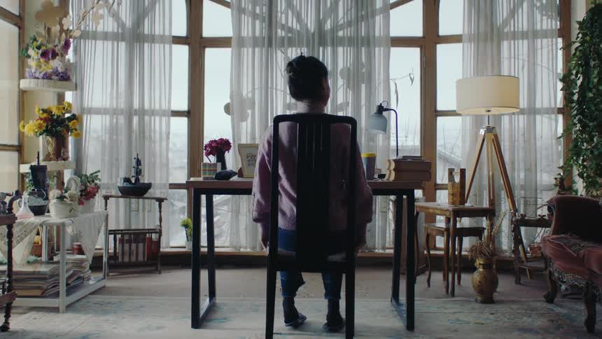 A woman in a cozy light filled room with vintage decor as she leans back in a chair near a desk framed by large arched windows adorned with sheer curtains
