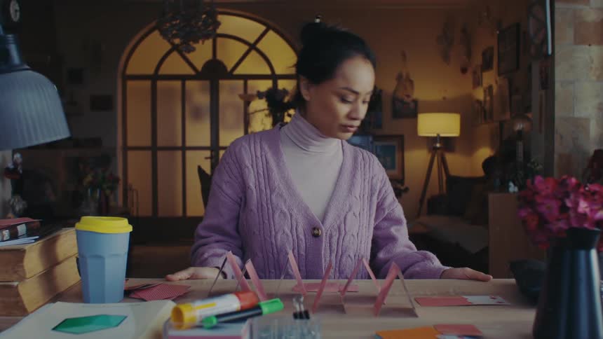 A person is sitting at a desk in a cozy warmly lit room constructing a house of cards surrounded by various stationery items books and a lamp