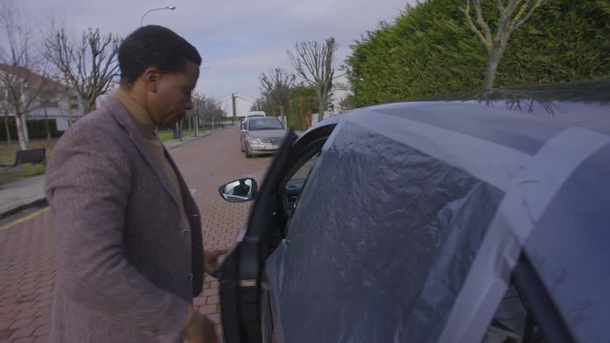 Man running his hand through a cover covering a car window and then he picks up the tape and gets into the car