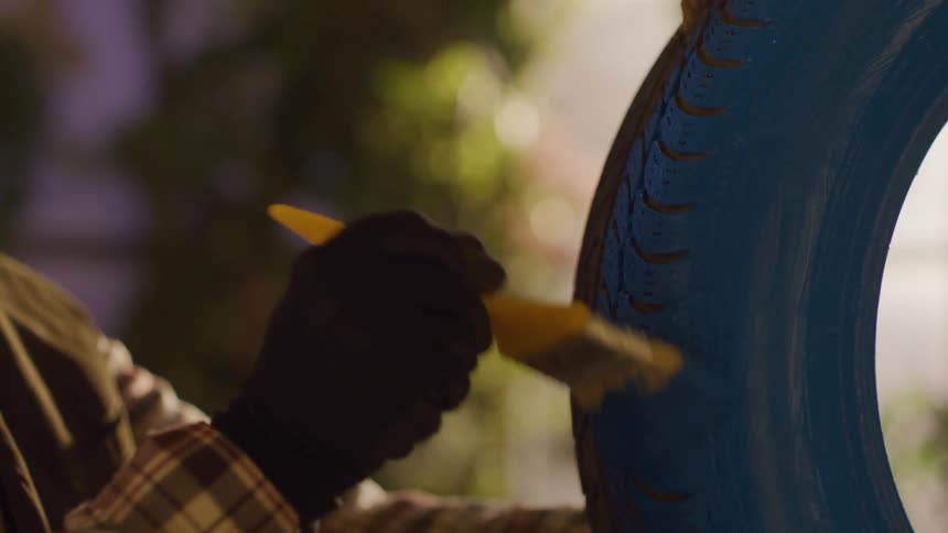Man paints a tire hanging on a rope blue while indoors during the daytime
