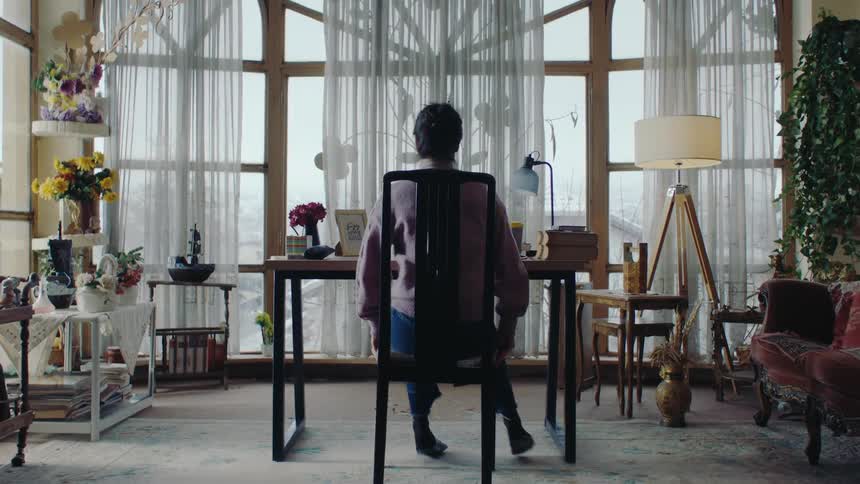 A woman in a lilac sweater and jeans stands checking a chair then sits at a desk in a room with large windows and various decorative items