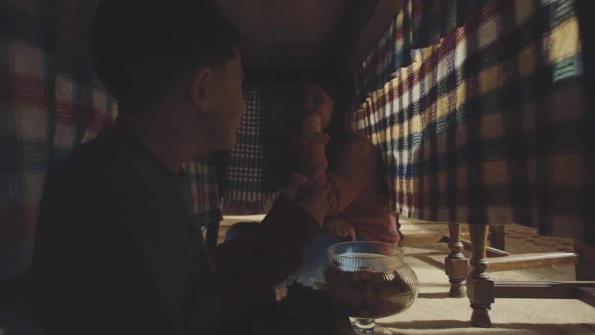 Siblings are sitting under a table covered with a checkered tablecloth eating cookies from a glass bowl