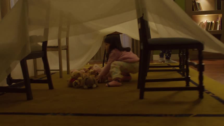 Two children covering four chairs with a sheet with a little girl sitting in the center forming a hut