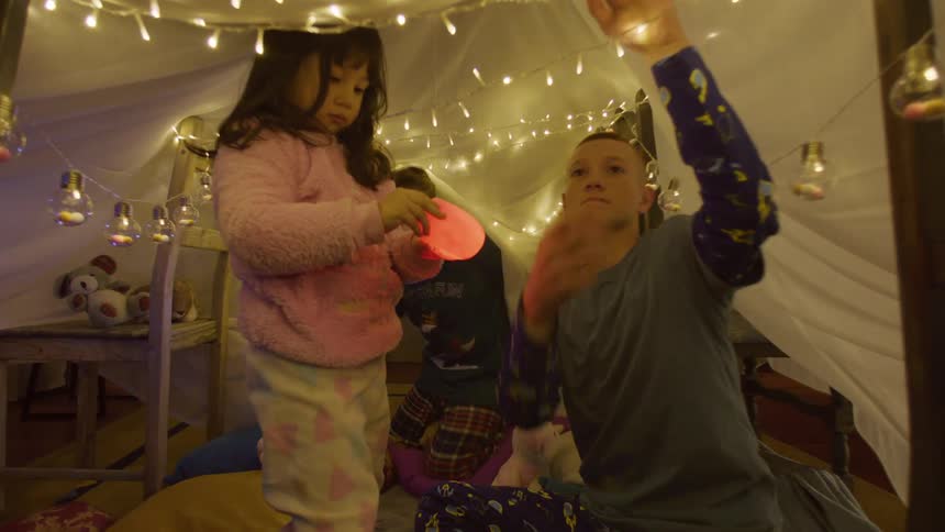 Three small children playing under a blanket with chairs around and lights