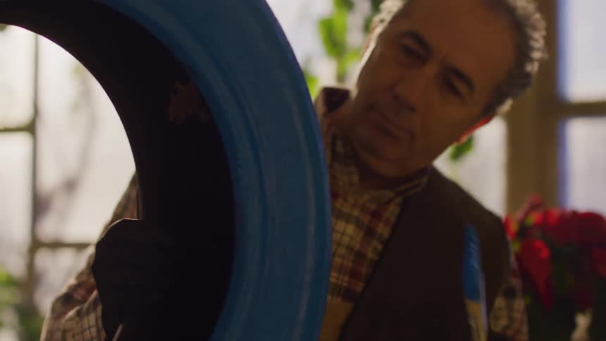 Middleaged man wearing a plaid shirt and dark vest painting a blue tire with a yellow paintbrush