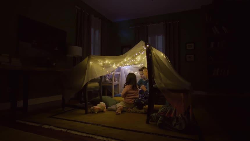 A young girl sits inside a cozy homemade blanket fort illuminated by warm fairy lights accompanied by two boys one of whom is holding a flashlight adding to the sense of adventure and childhood wonder in the dimly lit room