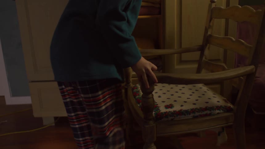A young kid in a teal sweater steps onto a chair to reach into a wooden wardrobe to place or retrieve folded blankets or towels