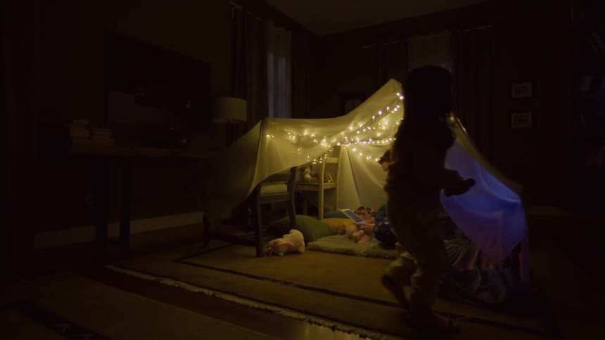 Child holding a flashlight in a dark room illuminating a makeshift hut made of sheets decorated with twinkling lights
