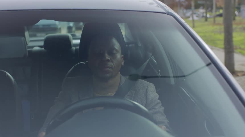 A man wearing a blazer is driving a car seen through the windshield on a city street