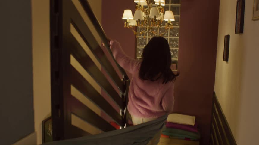 Little girl wearing a pink coat and light pants walking down a staircase carrying a blanket