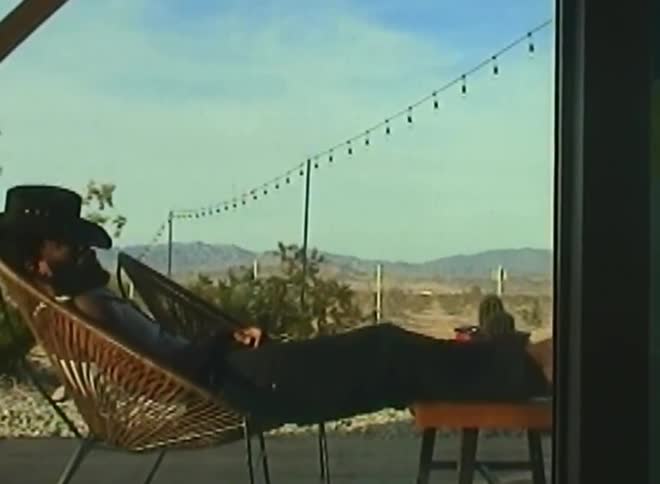 A person wearing a cowboy hat is lounging in a wicker chair with legs propped up on a small table set against a backdrop of desert landscape and string lights