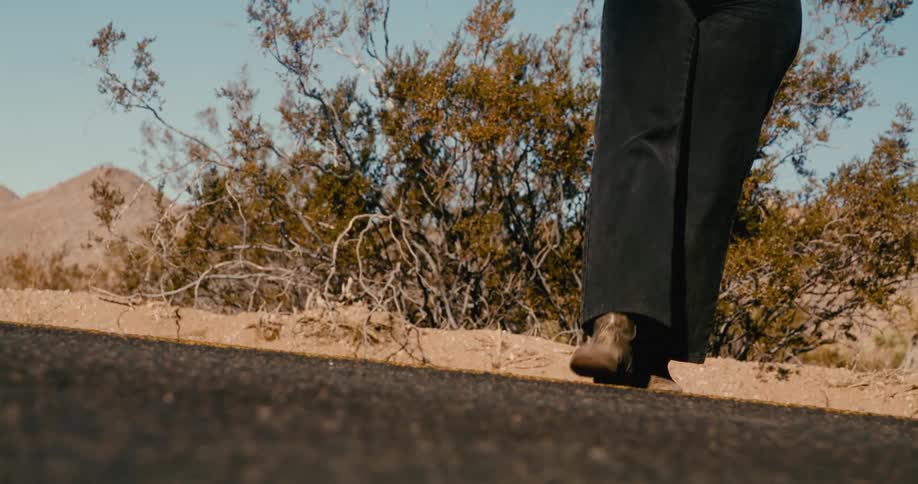 A woman wearing dark pants and tan boots walks along a dark curb in a desert landscape with dry brush and mountains in the background