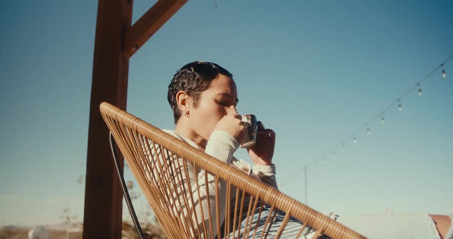 A person is sitting in a wicker chair under a wooden structure drinking from a cup with string lights and a clear blue sky in the background
