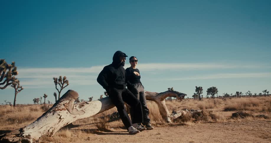 Woman and a man lean against a fallen log in a desert area with sparse vegetation