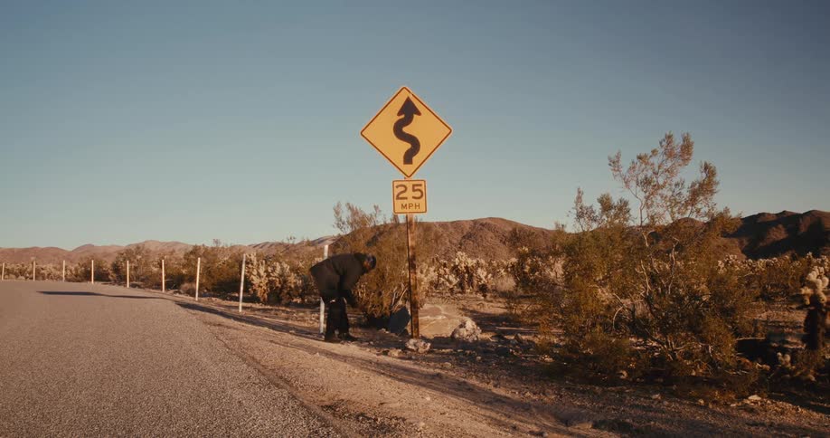 Man walking along the side of a road and leaning against a sign in a desert location