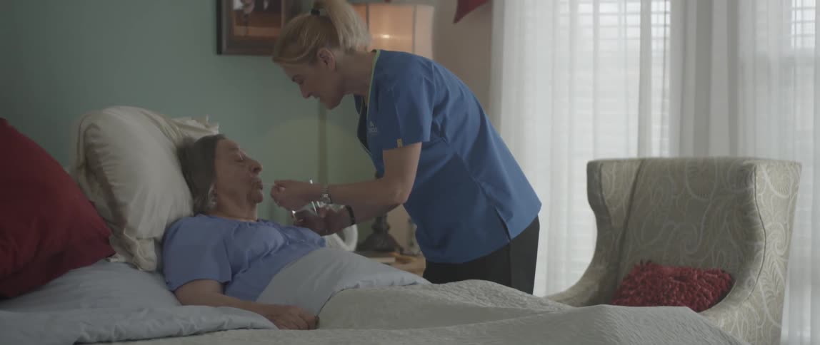 A healthcare worker offers a drink of water to an elderly woman laying in bed