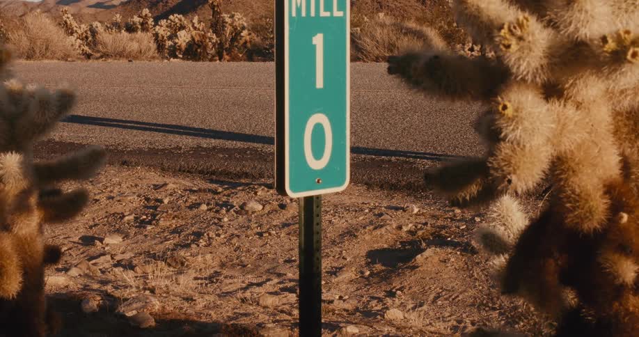 A green mile marker sign with the number 10 stands in a desert landscape with a person walking on the road in the background and cacti on either side