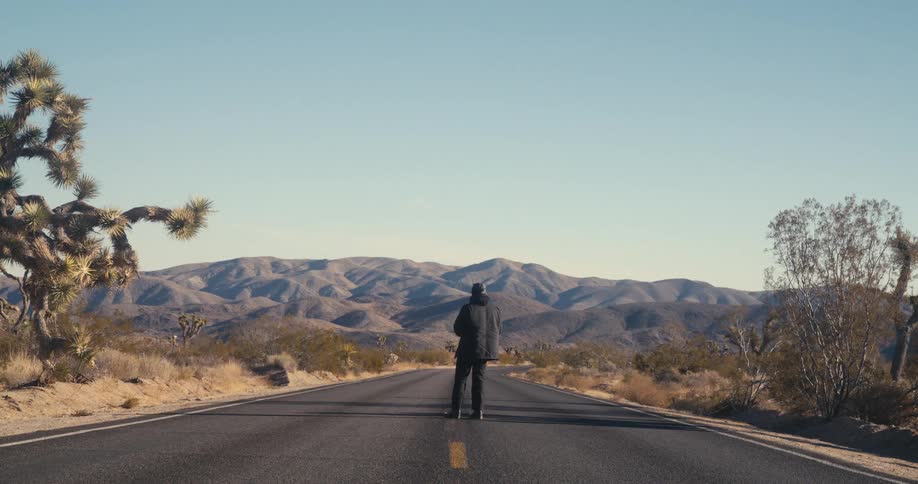 Man crossing a highway in the desert pauses to look towards the mountains