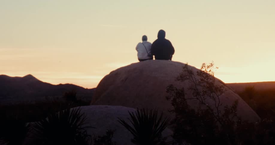 Two people are back to back and sitting on top of a rock looking forward where there is a white sky with sunlight coming from the horizon