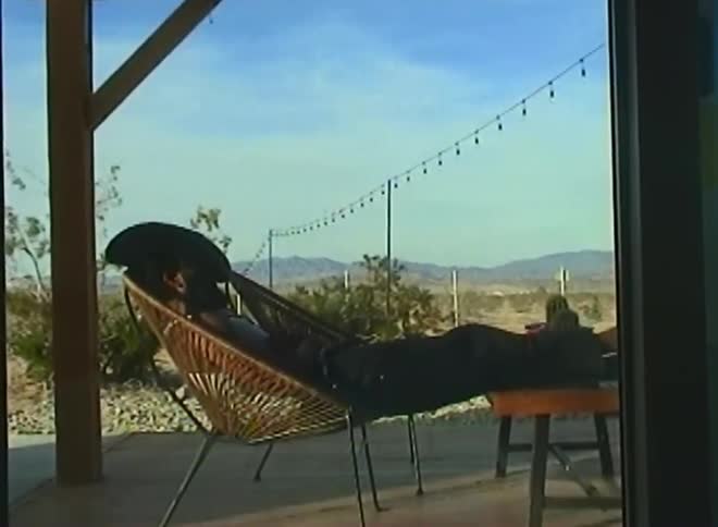 A man in a hat sleeping in a chair that is on a porch of a house and then he gets up