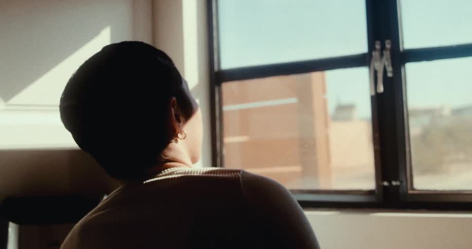 Woman raises the blinds to let the sunlight in just as a semi truck passes on the nearby highway