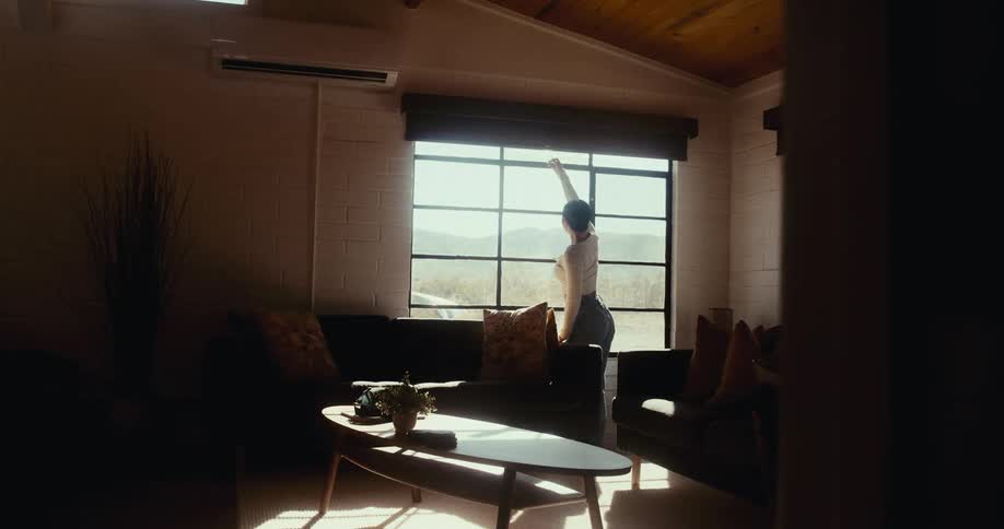 A person is standing by a large window in a dimly lit living room adjusting the blinds with sunlight streaming in and casting shadows on the furniture