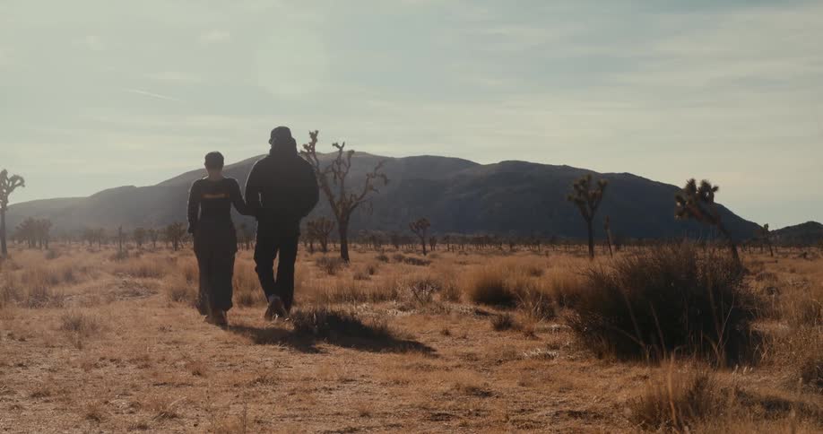 Two people are walking through a dry desert landscape with sparse joshua trees and mountains in the background under a partly cloudy sky
