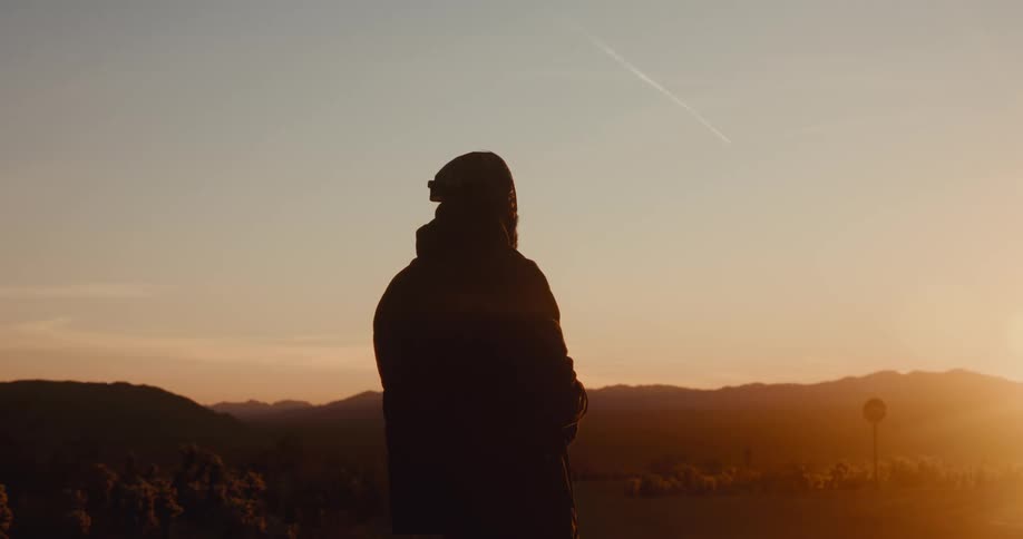 The silhouette of a person in long clothes and he is standing looking forward where there is a sunrise on the horizon