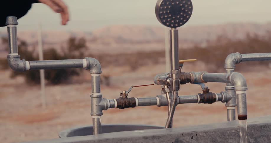 A person in a black garment reaches for an outdoor shower fixture made of industrial pipes with water spraying from the showerhead against a blurred dry landscape backdrop