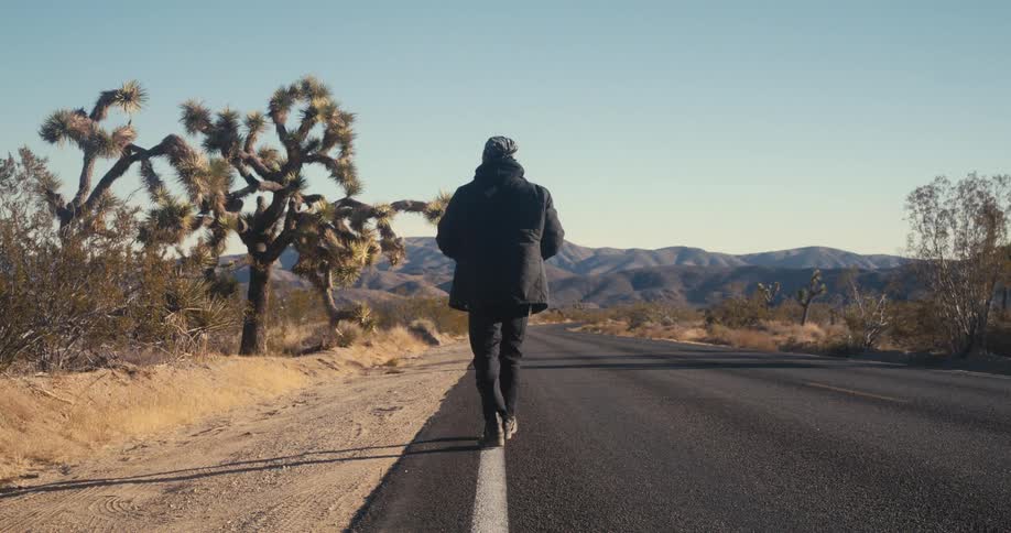 Man in black walking along a paved road in a desert environment with mountains on the horizon