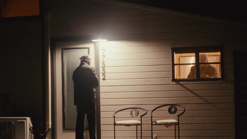 Man entering a house at night and another man messing with a camera seen through a window