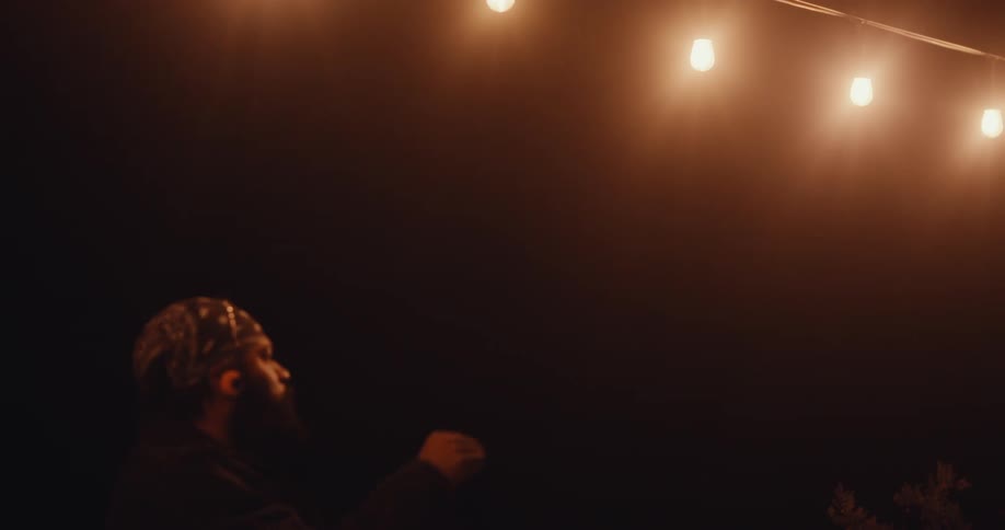A person wearing a bandana is dancing outdoors at night under a string of illuminated light bulbs