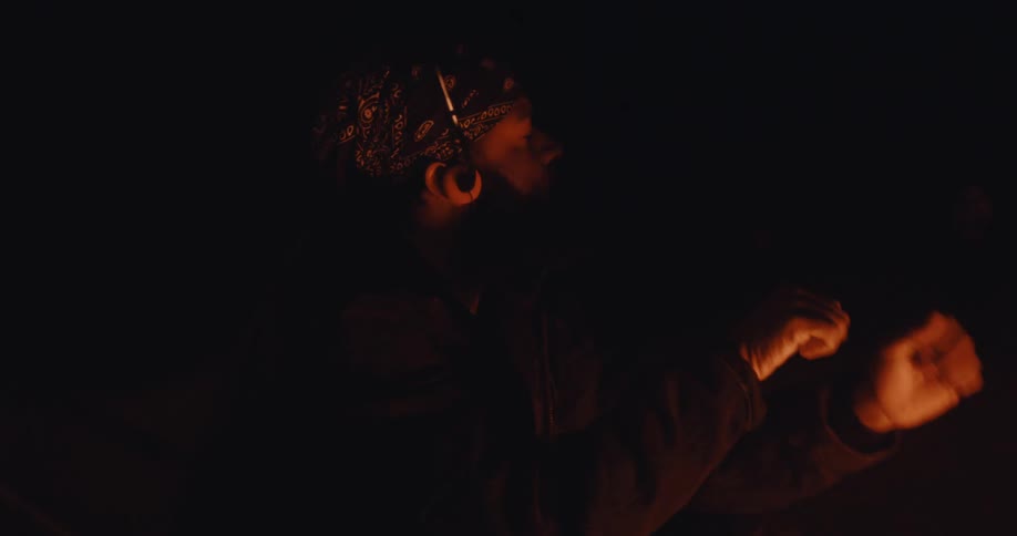 Man in bandana and dark jacket sits in the dark lit by orange glow of fire enjoying music with headphones