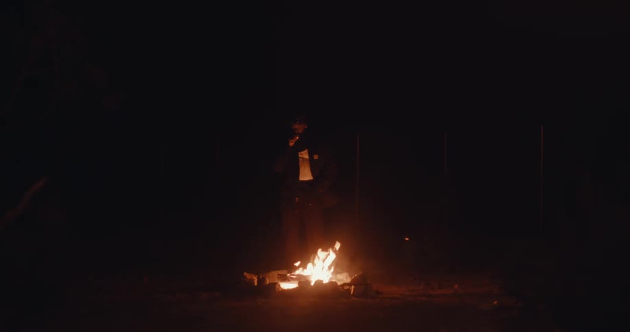A man walks to a fire on the ground that is lighting up the dark surroundings and he stands in front of it