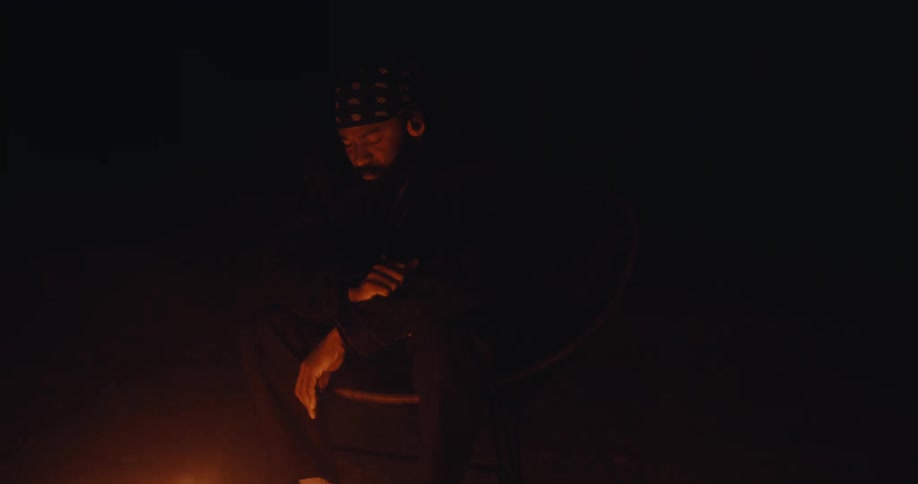 A man seated on the ground in a dark environment in front of a fire that is illuminating him and he makes gestures with his hand as if he is listening to music
