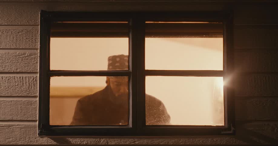 A mysterious man wearing a bandana and dark clothing standing outside a window and filming or observing with a handheld camera