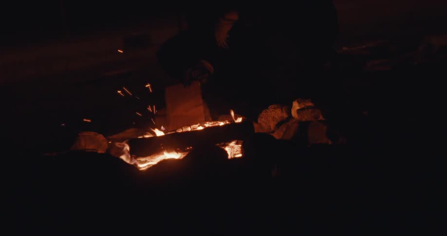 Person wearing gloves adds wood to a campfire at night