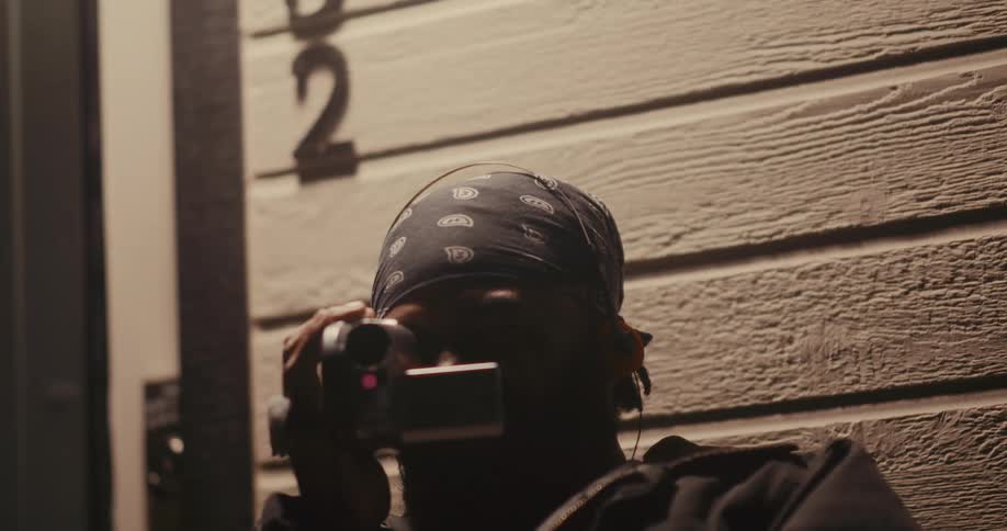 A bearded man wearing a bandana leaning against the wooden wall pulls out a handheld camera and begins filming