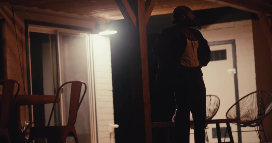 A man standing under the dim light of a porch at night wearing a bandana jacket and tank top gazing upwards with a contemplative or intense expression while an outdoor seating area and a sliding glass door add to the quiet moody atmosphere