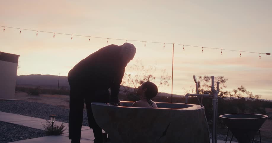 A man approaches a woman who is in an outdoor jacuzzi and he puts a hat on her head