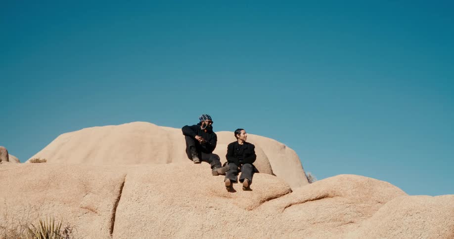 Two people wearing dark clothes are sitting on large lightcolored rocks in the desert under a cloudless blue sky