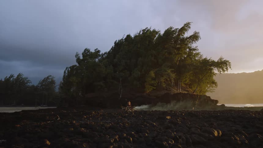 A shirtless man stands near dense trees starting a bonfire as smoke drifts to the right