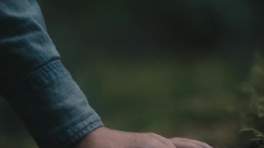 A blonde womans hand wearing a denim jacket and sporting long dark nails rests gently on a mossy rock