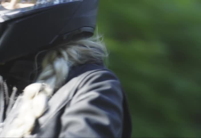 A person with long blond hair wearing a black helmet and a jacket riding a motorcycle