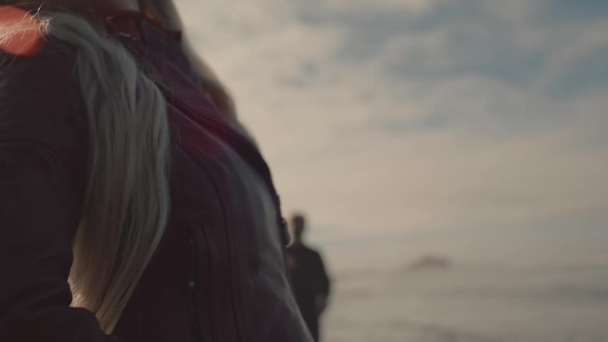 A person in a black jacket and long blond hair standing and behind there is another person