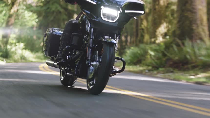 A person is riding a black motorcycle through a winding road in a forest with the front wheel and engine visible in motion