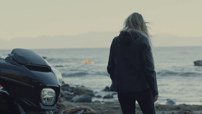 An older woman with shoulderlength silver hair in a leather jacket walks away from a parked black motorcycle toward the coastal water as small waves roll ashore