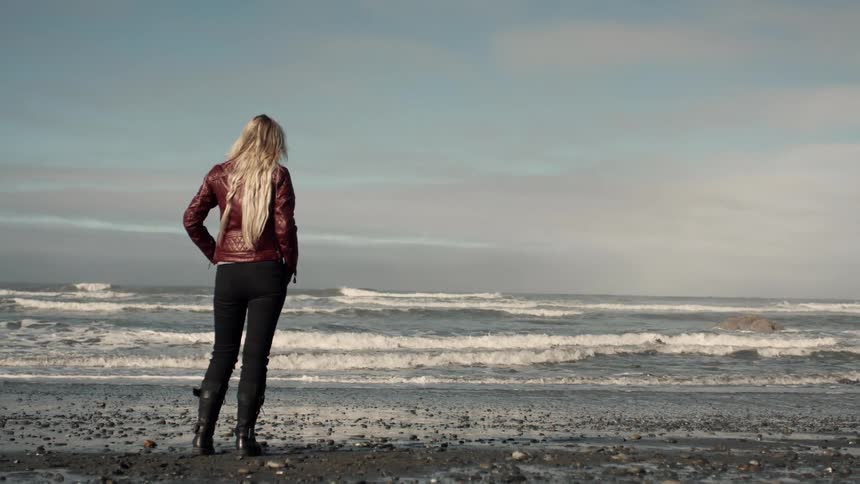 Woman in red jacket and black boots stands with her back on the beach looking at the rough sea with waves crashing on the shore
