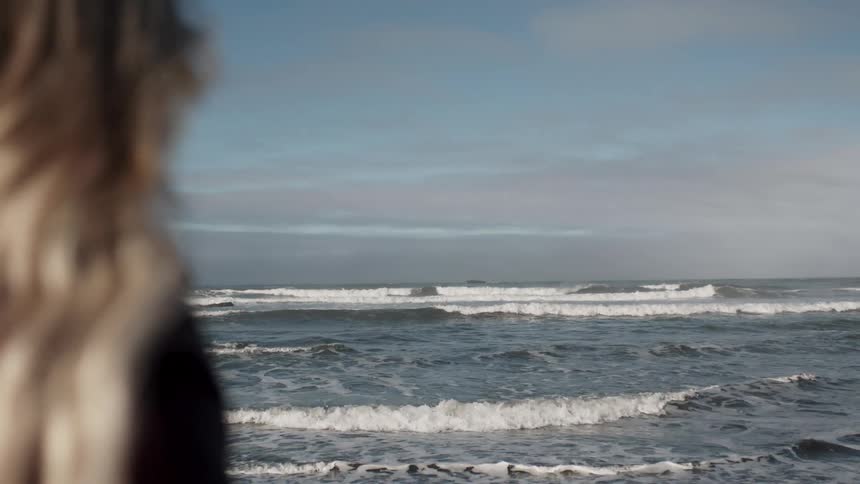 A woman with long wavy blonde hair is standing on a beach looking out at the ocean waves under a clear blue sky